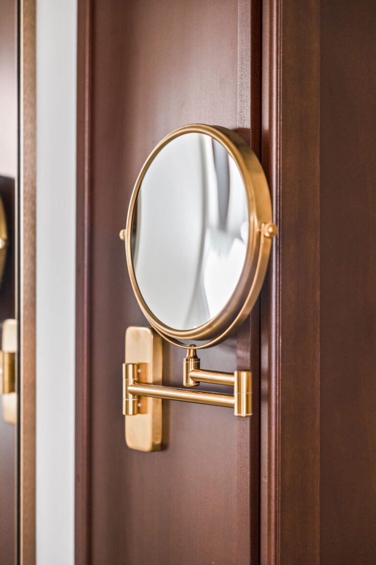 A Carmel, IN Bathroom Renovation picture showing a magnifying mirror in matte brass attached to the rich wood staining vanity taller cabinet.