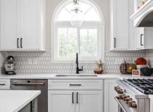 An arched window by a kitchen sink and white cabinets.