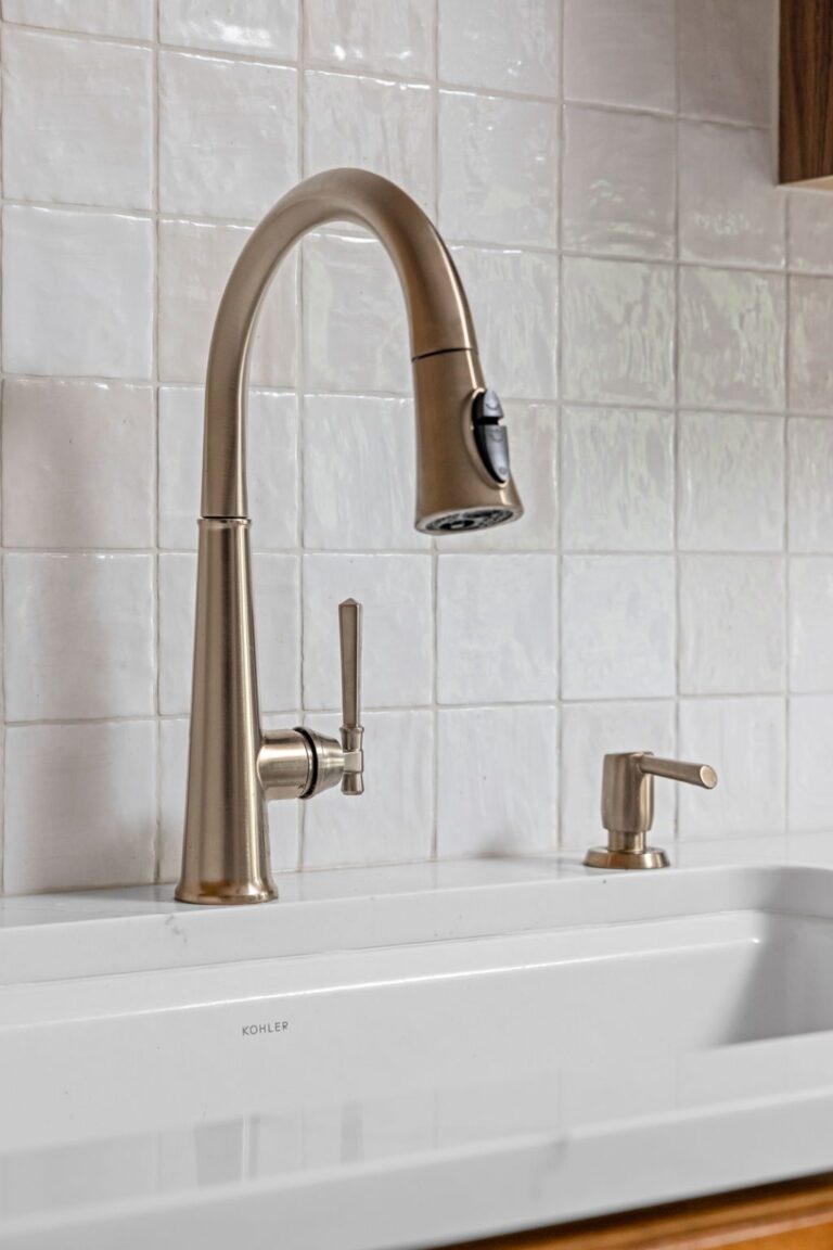 Brushed brass kitchen faucet and soap dispenser mounted at a farmhouse sink with white tile backsplash.