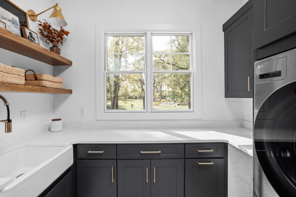 an organized laundry room remodel in carmel indiana with custom cabinetry in gray and custom cabinetry inserts of trash drawers, folding trays, pet feeding station drawer, soaking sink, drawer drying racks, white countertops and a mudroom incorporated with broom storage