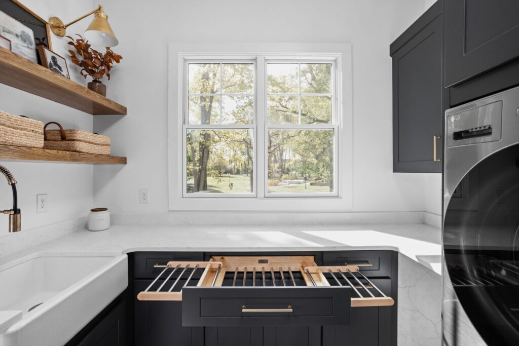 an organized laundry room remodel in carmel indiana with custom cabinetry in gray and custom cabinetry inserts of trash drawers, folding trays, pet feeding station drawer, soaking sink, drawer drying racks, white countertops and a mudroom incorporated with broom storage