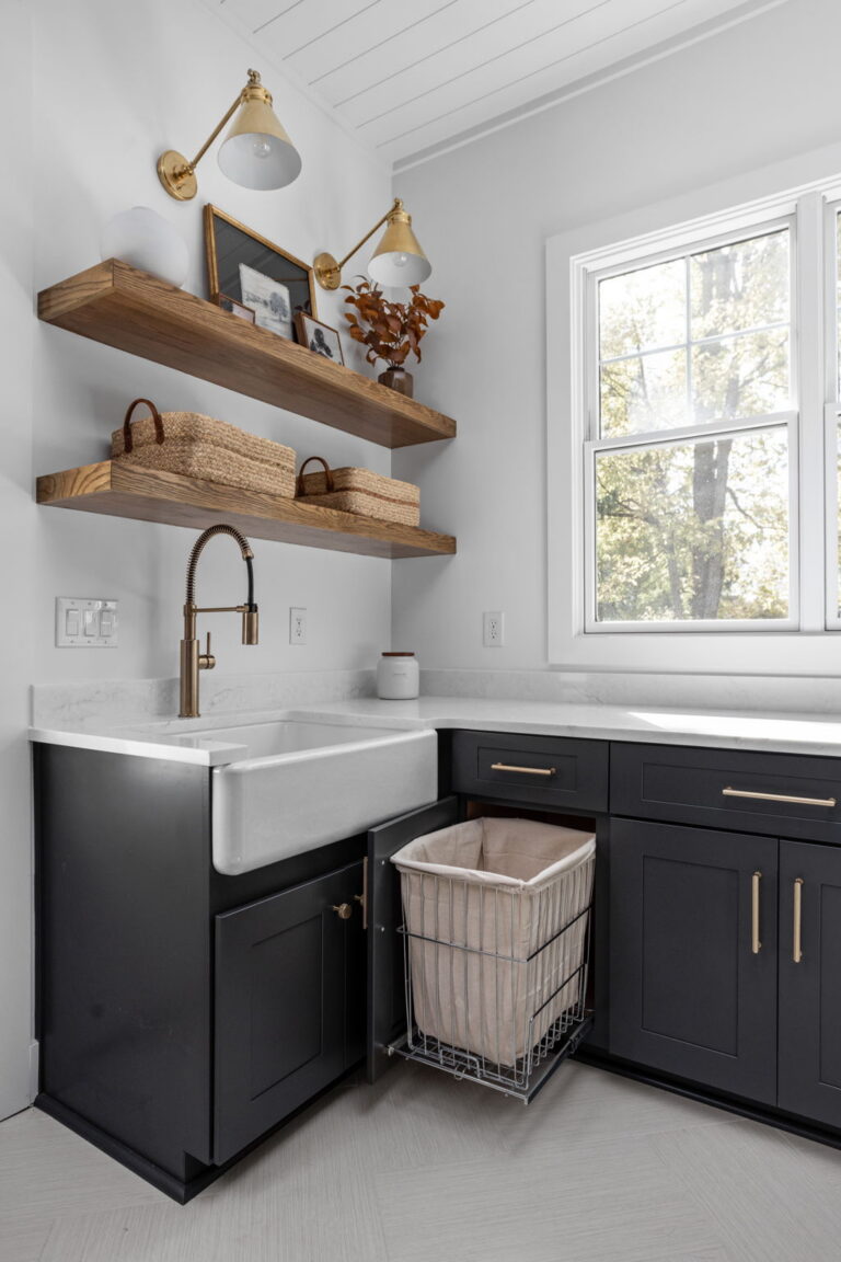 an organized laundry room remodel in carmel indiana with custom cabinetry in gray and custom cabinetry inserts of trash drawers, folding trays, pet feeding station drawer, soaking sink, drawer drying racks, white countertops and a mudroom incorporated with broom storage and floating wood shelves