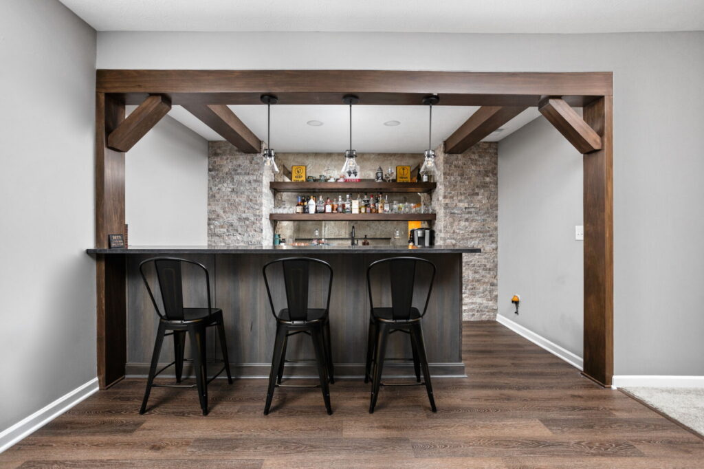 Basement wet bar with dark wood beams, gray cabinetry, bar seating, and stacked stone in Westfield IN