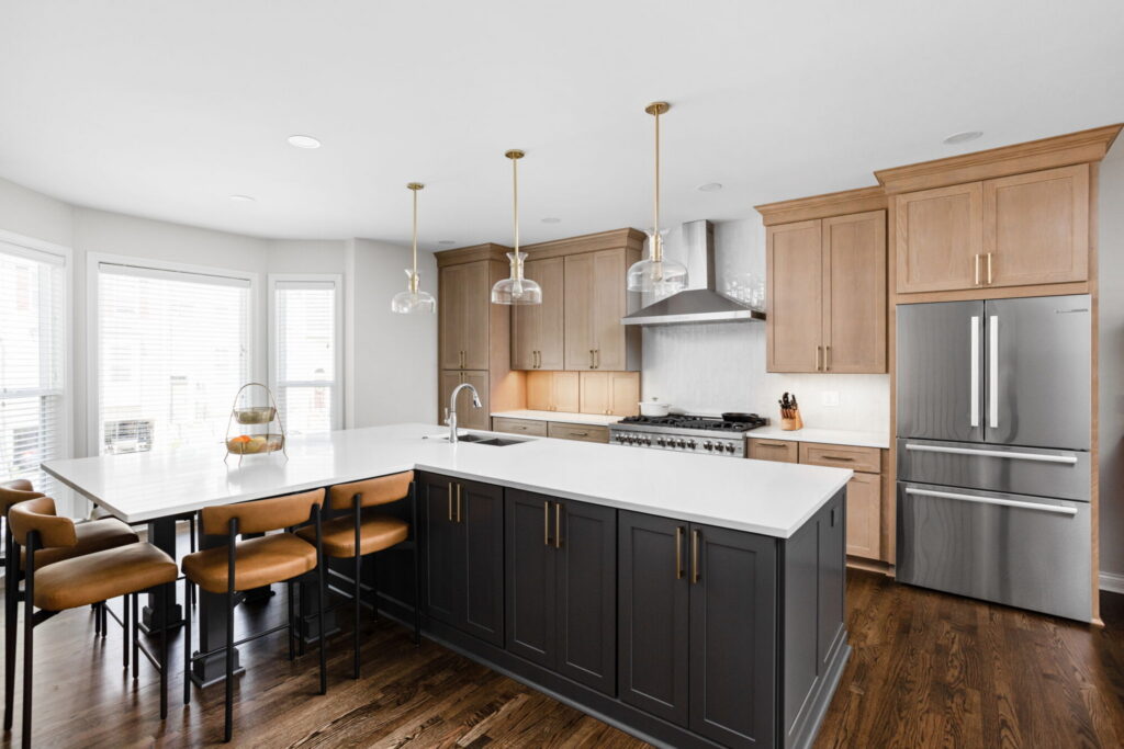 High end kitchen remodel Carmel featuring a large seating island, white countertops, light stained wood cabinets, brass pendant lighting, and stainless appliances.