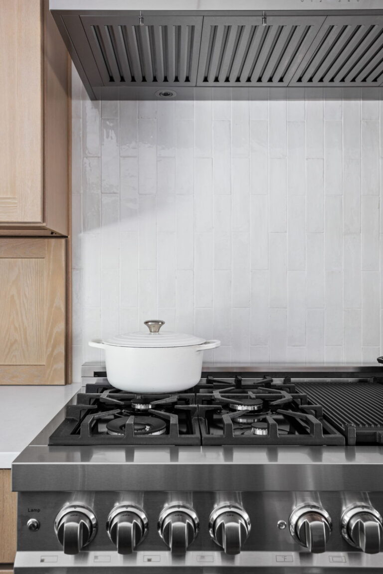 High end kitchen remodel Carmel highlighting a professional style gas range, white tile backsplash, and stainless vent hood beneath light wood cabinetry.
