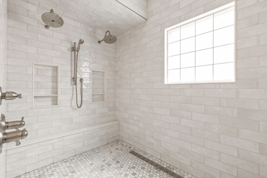Finished primary bathroom shower in a Carmel home with white wall tile, dual shower heads, recessed niches, glass block window, and linear drain