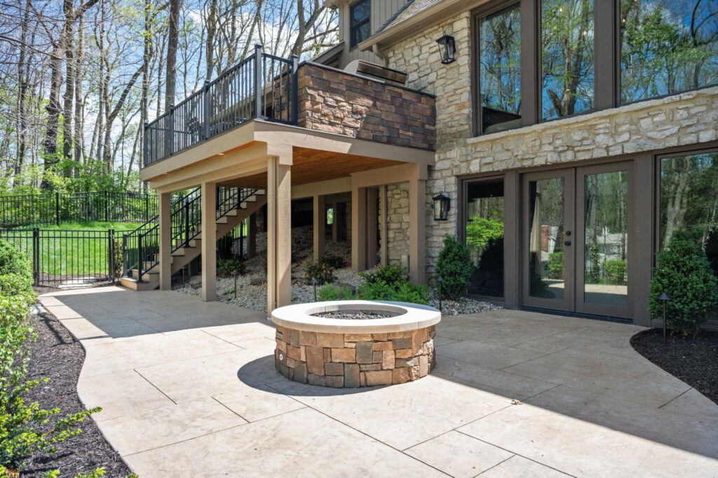 Stamped concrete patio in Carmel with gas firepit, Trex deck above, black railing, stone exterior, and updated basement doors.