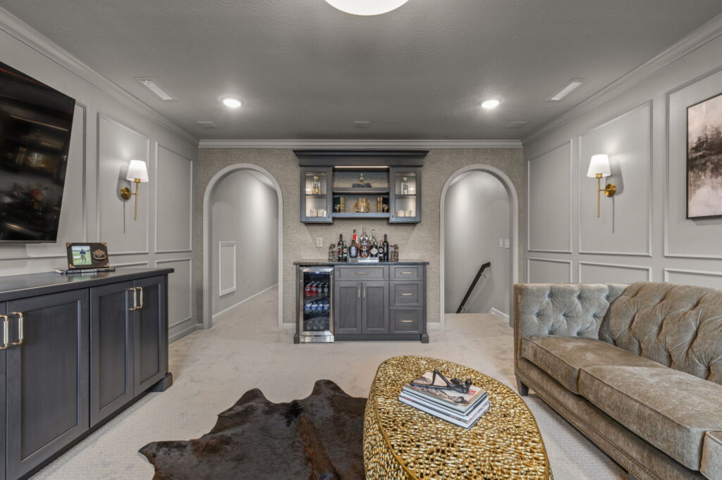 Custom home bar remodel in Fishers, Indiana showing the finished lounge with dry bar, TV storage cabinetry, arched openings, layered lighting, and seating area.