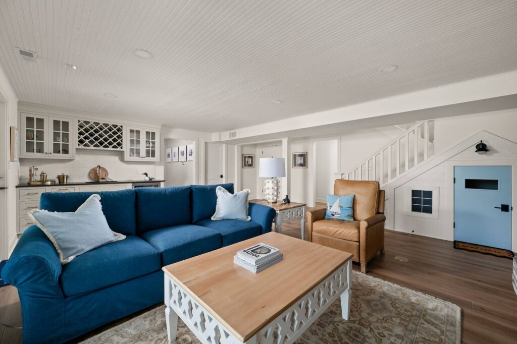 Lower-level living room in the Nantucket Bay whole-house remodel with bar, blue sofa, beadboard ceiling and under-stair playroom door.