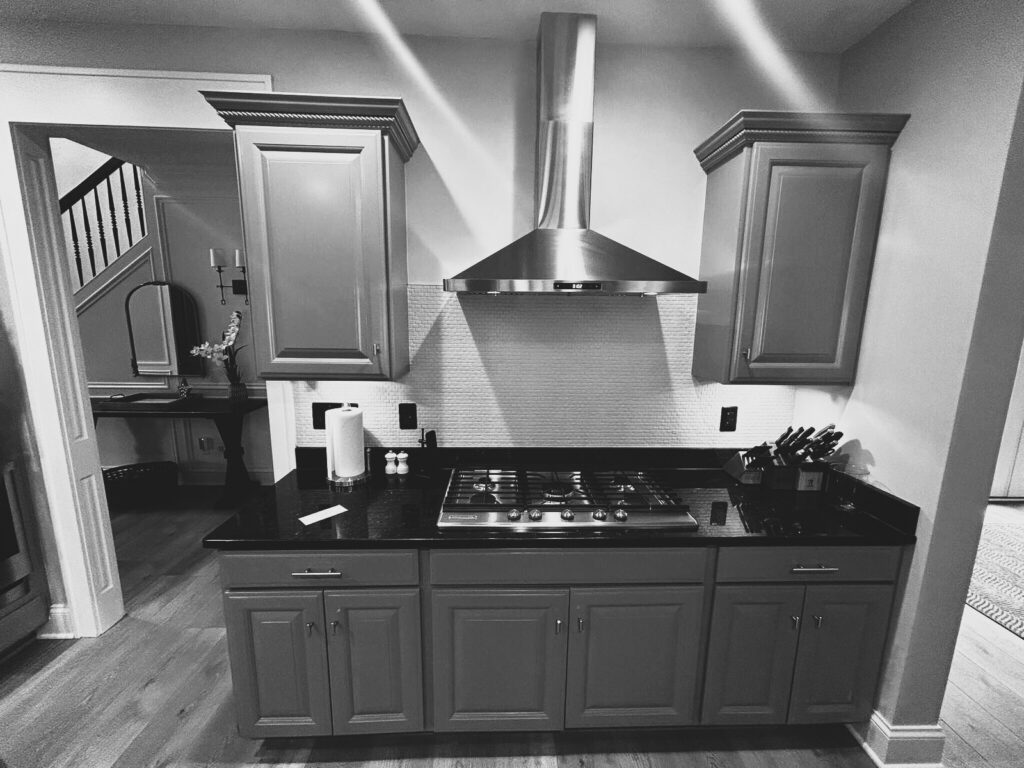 Before: Carmel kitchen cooktop wall with gray cabinets, dark countertop and stainless vent hood.