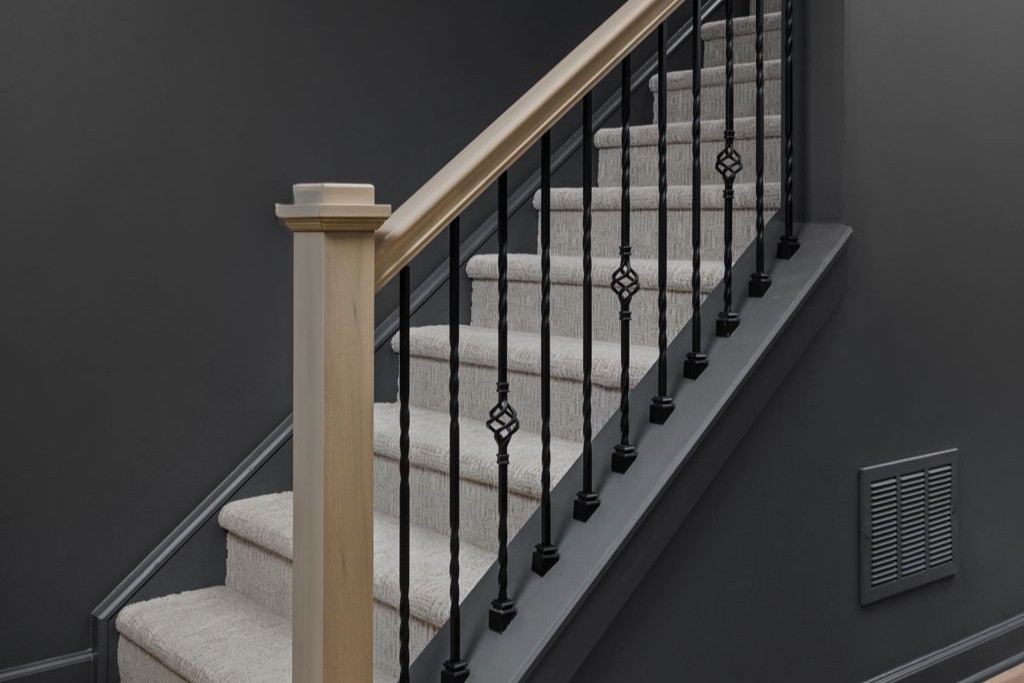 Carpeted basement stairs with a light wood handrail and newel post, black metal balusters and dark walls.