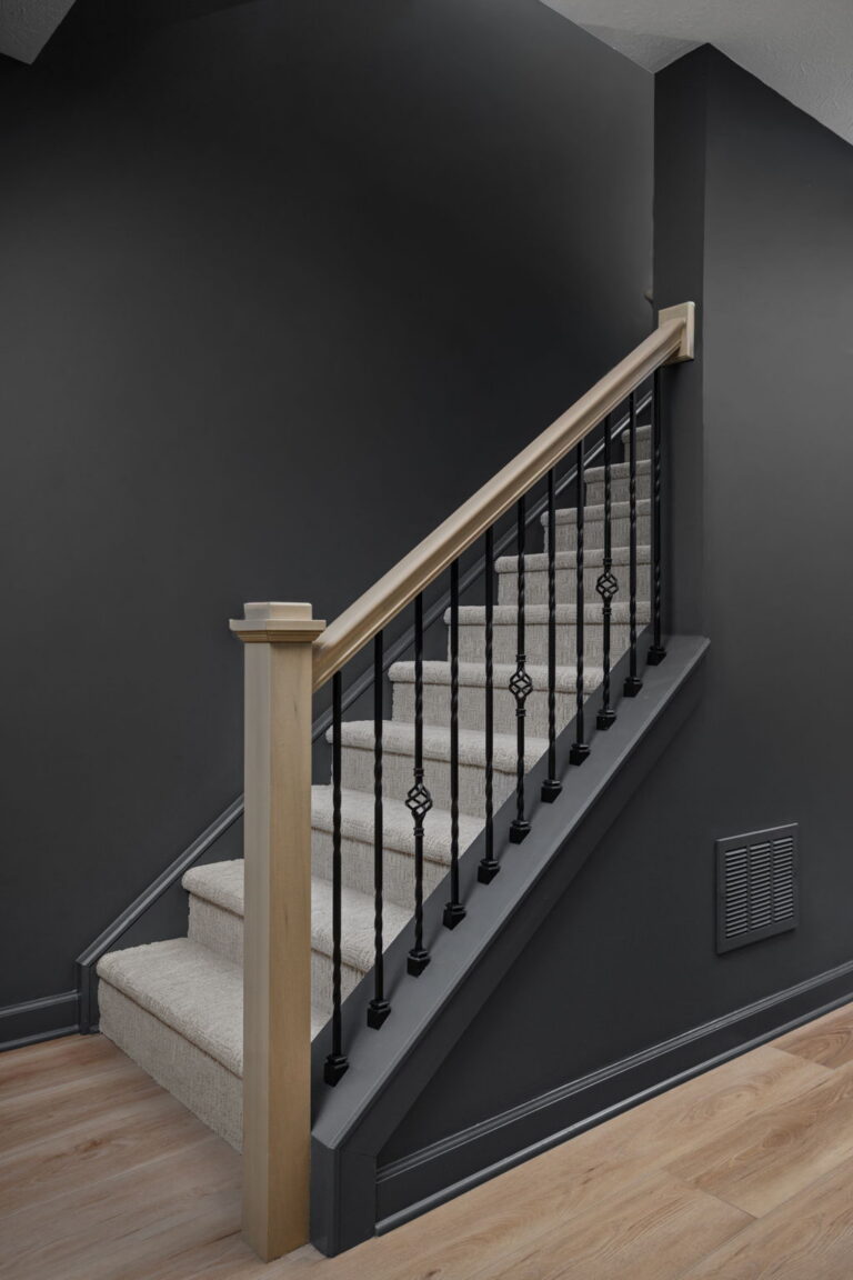 Carpeted basement stairs with a light wood handrail and newel post, black metal balusters and dark walls.