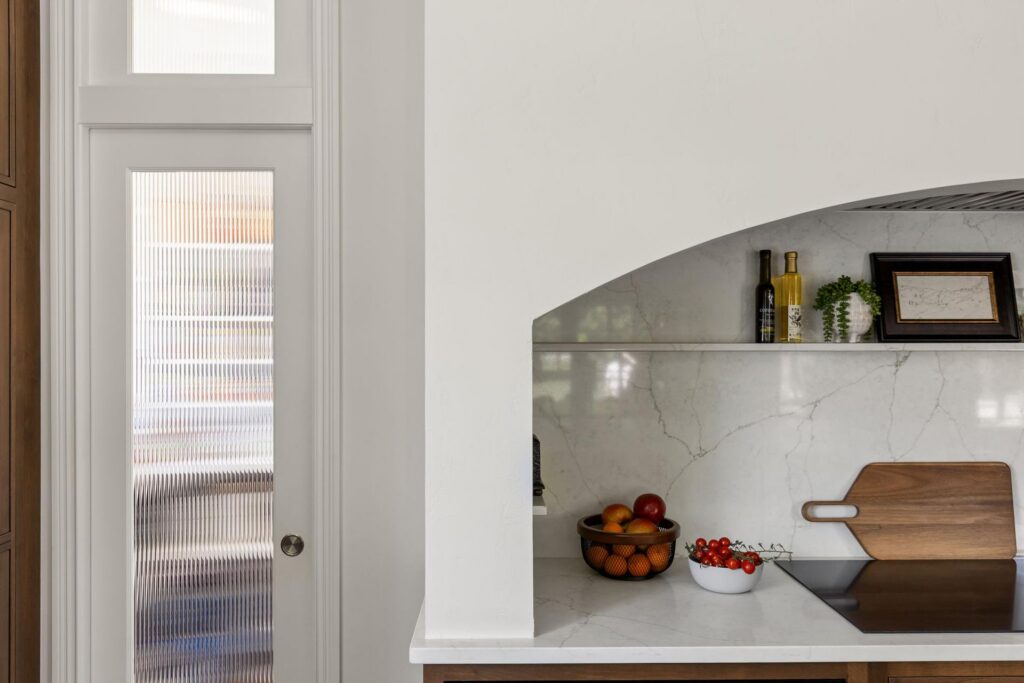 White arched kitchen hood with a veined quartz backsplash and recessed shelves beside a reeded-glass pantry door.