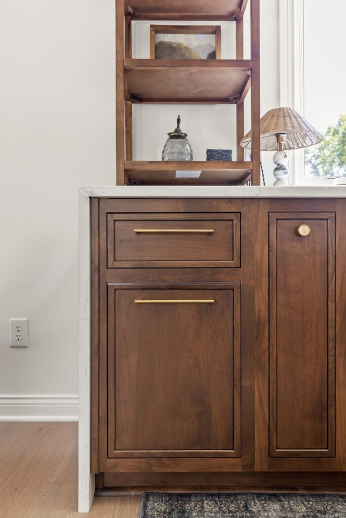 Inset wood cabinet doors and drawers with bronze-toned hardware beneath a quartz counter and open shelving beside the window.