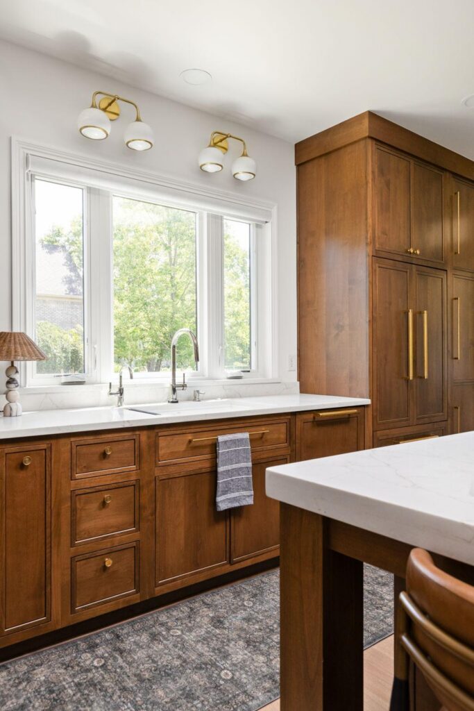 White kitchen sink beneath three windows, with a pull-down faucet, wood cabinetry and paneled refrigerator alongside.