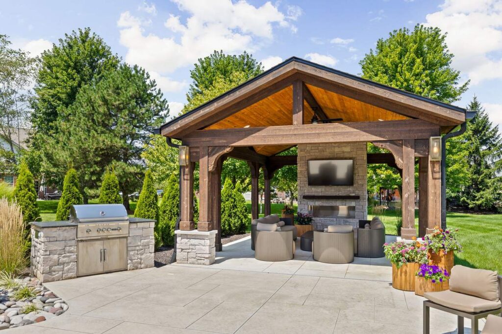 Backyard pavilion in Carmel with an open gable roof, stone fireplace and adjacent grill island.