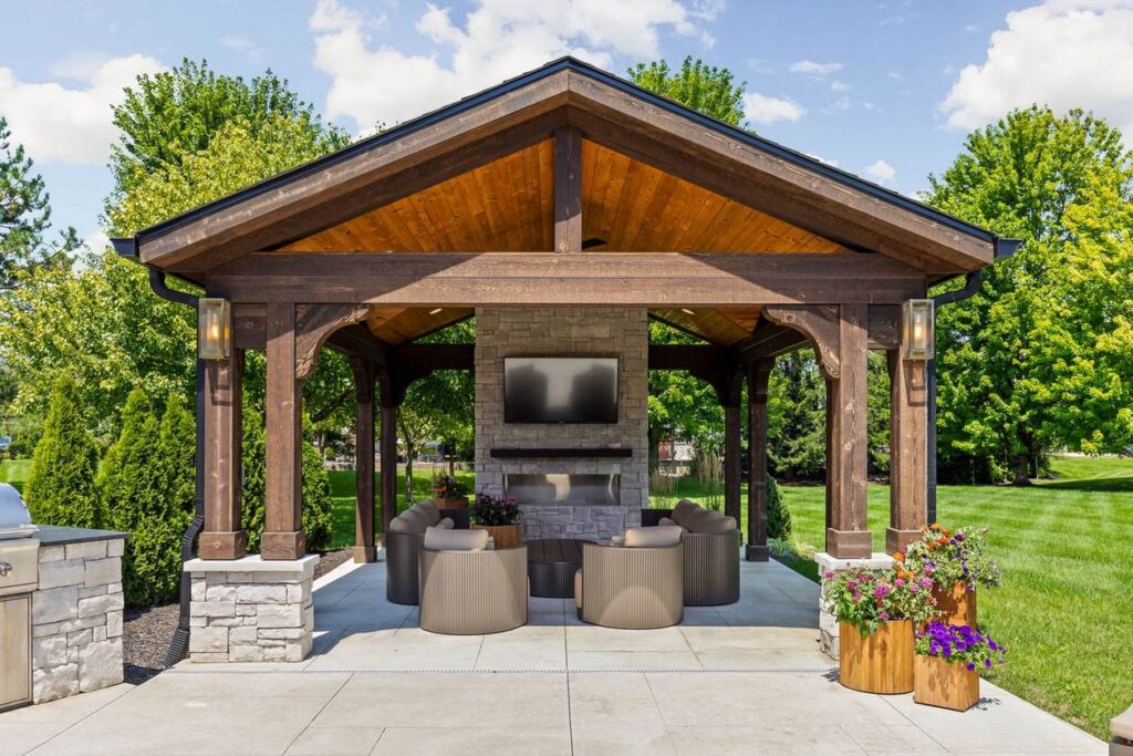 Front view of the backyard pavilion with dark wood posts, stone column bases, a fireplace and outdoor seating.