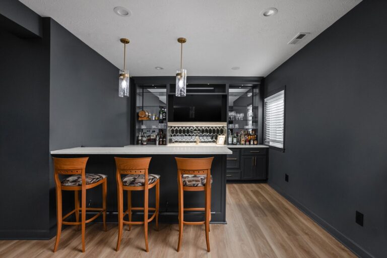 Finished basement bar in Noblesville with dark cabinetry, three wood stools, a light countertop and glass pendant lights.